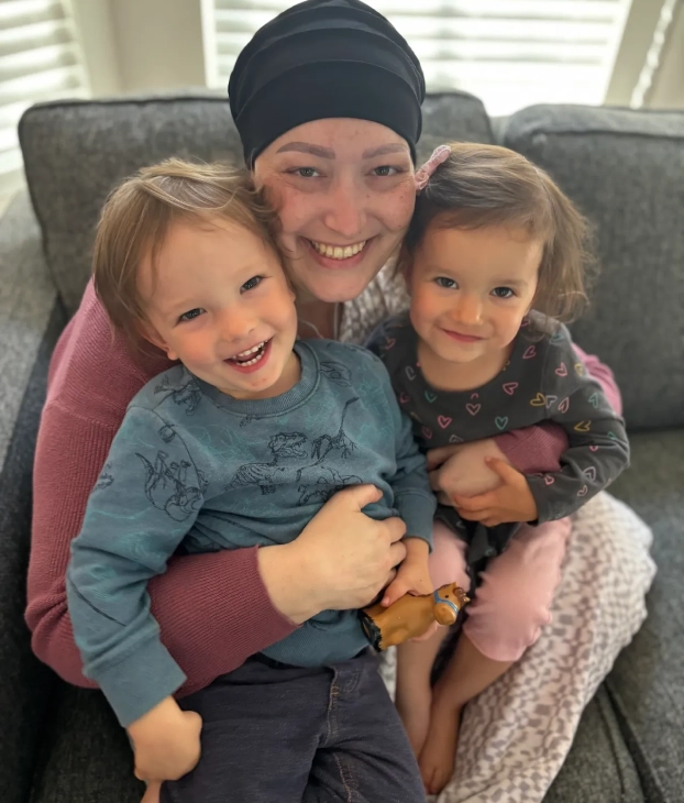 A smiling woman hugs two young children on a couch.